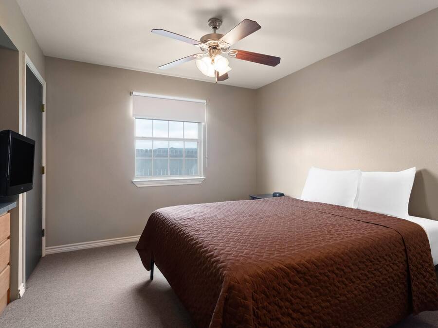 Un dormitorio con cama, ventilador de techo, ventana y televisor sobre un aparador.