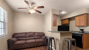 A compact living space with a brown leather sofa, a ceiling fan, a kitchen area with wooden cabinets, and a black refrigerator. - Eagle's Den Suites Carrizo Springs a Travelodge by Wyndham (Carrizo Springs)