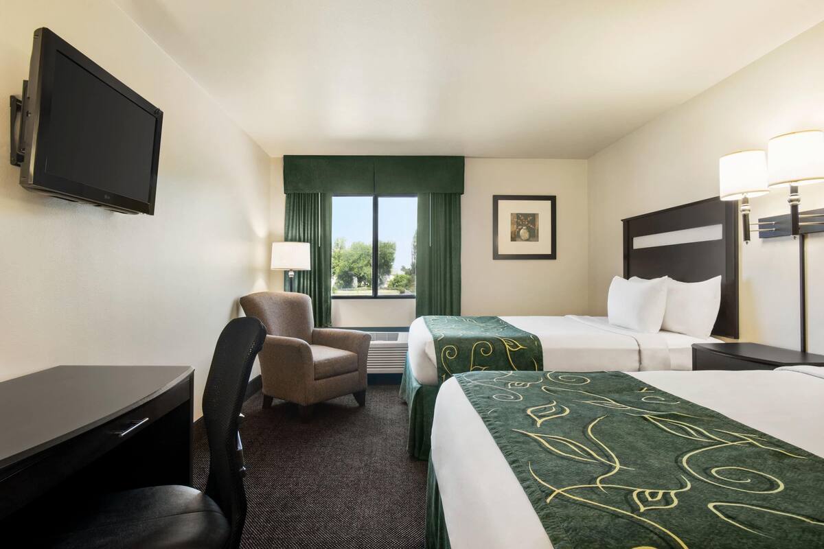 A hotel room with a desk, chair, two beds, a TV, a window with curtains, and a framed picture on the wall.