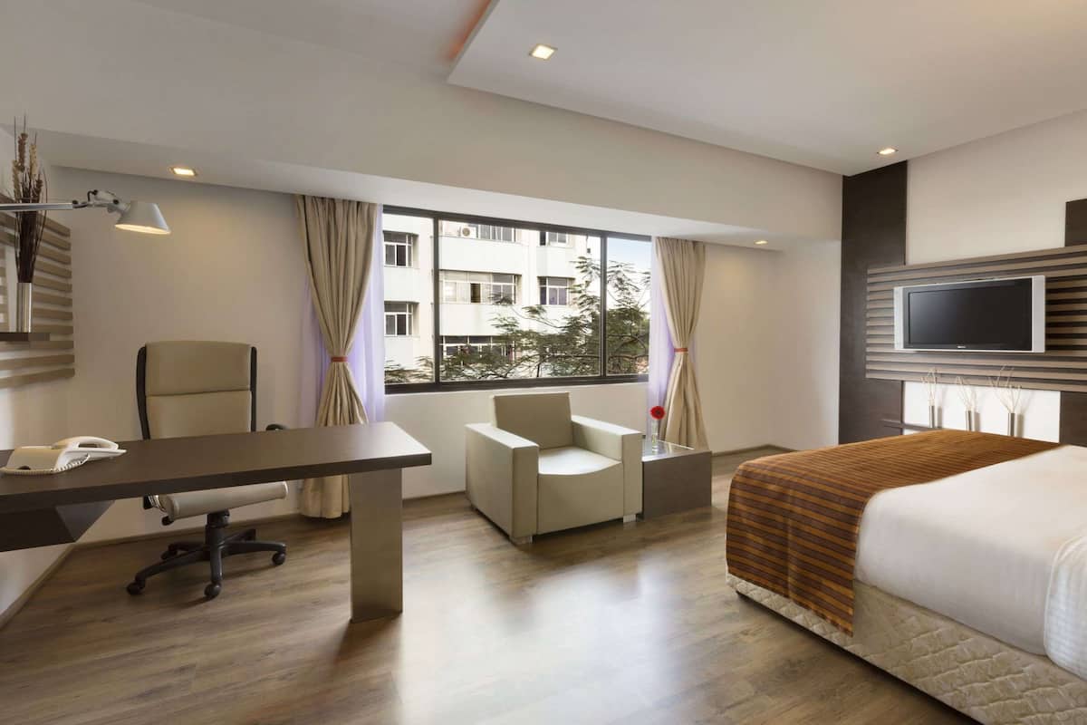 A hotel room with a bed, a desk with a phone, a chair, a TV, and a large window with curtains.