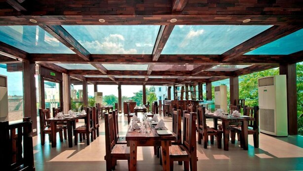 A spacious outdoor dining area with wooden tables and chairs, large glass windows, and a view of greenery.