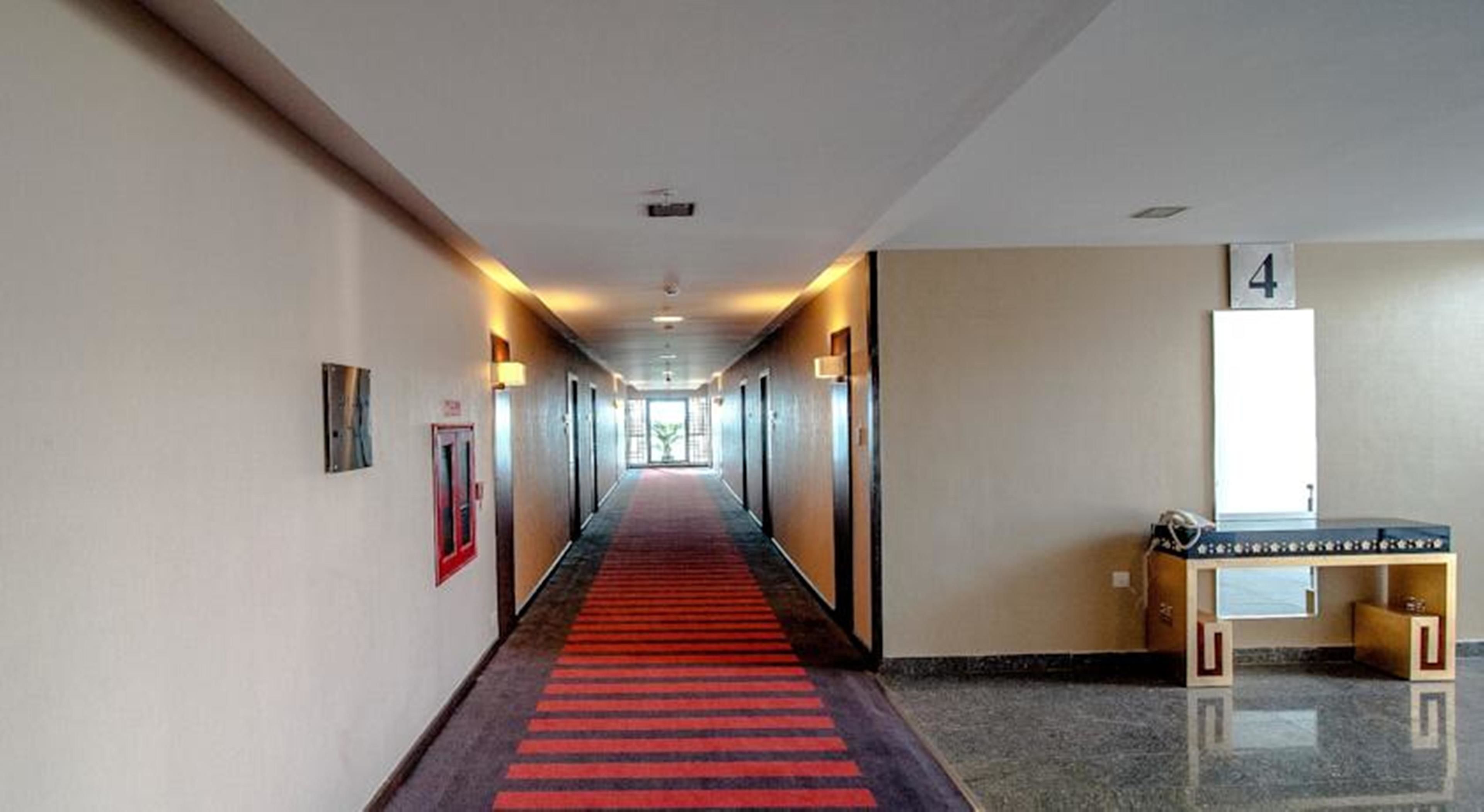 A hotel hallway with a red carpet, doors on both sides, and a desk with a computer on the right.