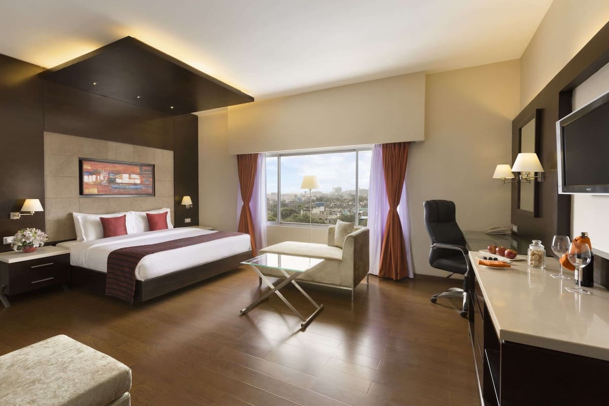 A modern hotel room with a bed, desk, chair, and a large window offering a city view.