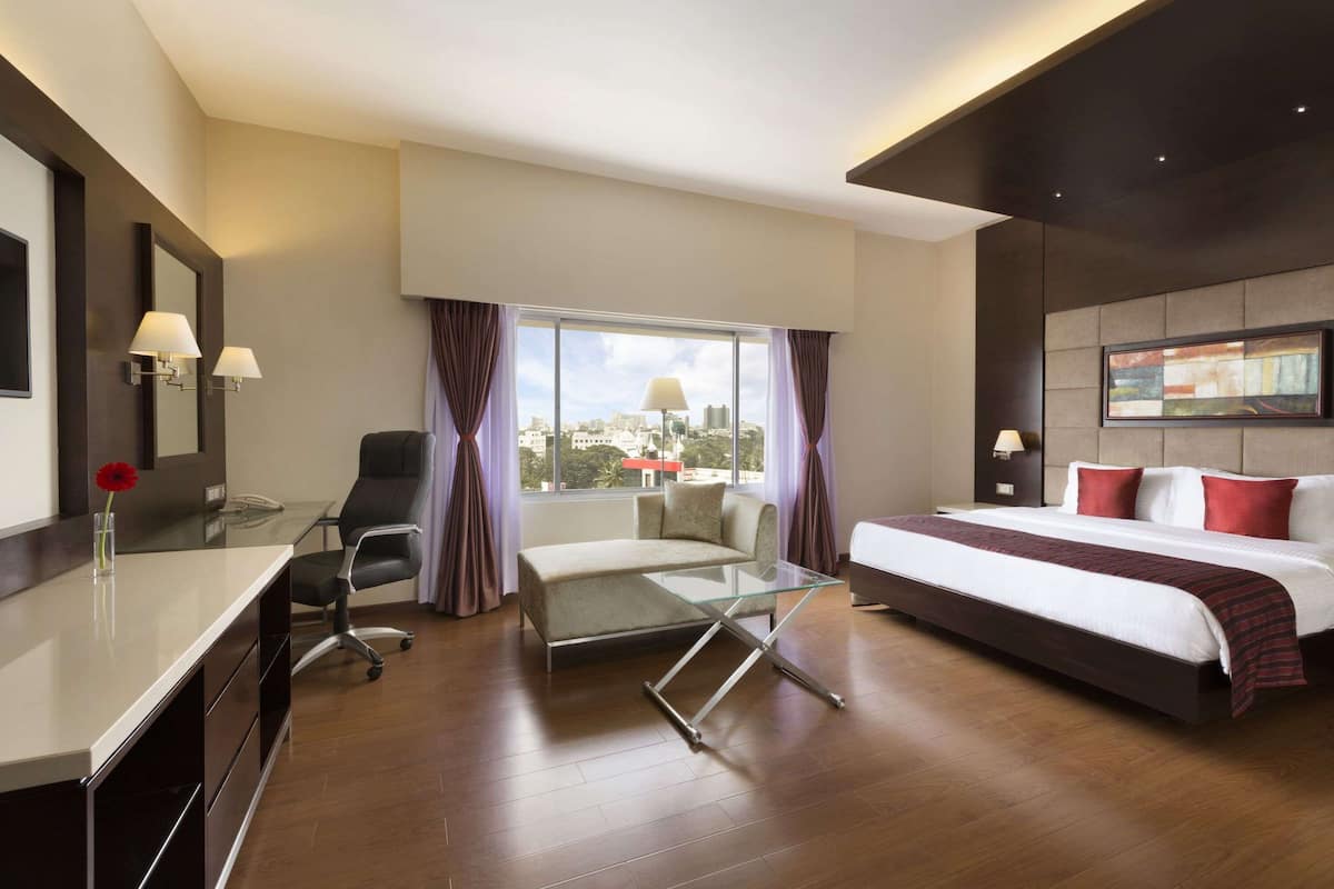 A modern hotel room with a glass desk, a sofa, a bed, and a city view.