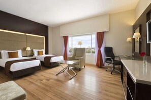 A hotel room with two beds, a desk, a chair, and a view of the city.