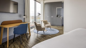 A hotel room with a bed, a desk, two chairs, a large window, and a kitchenette.