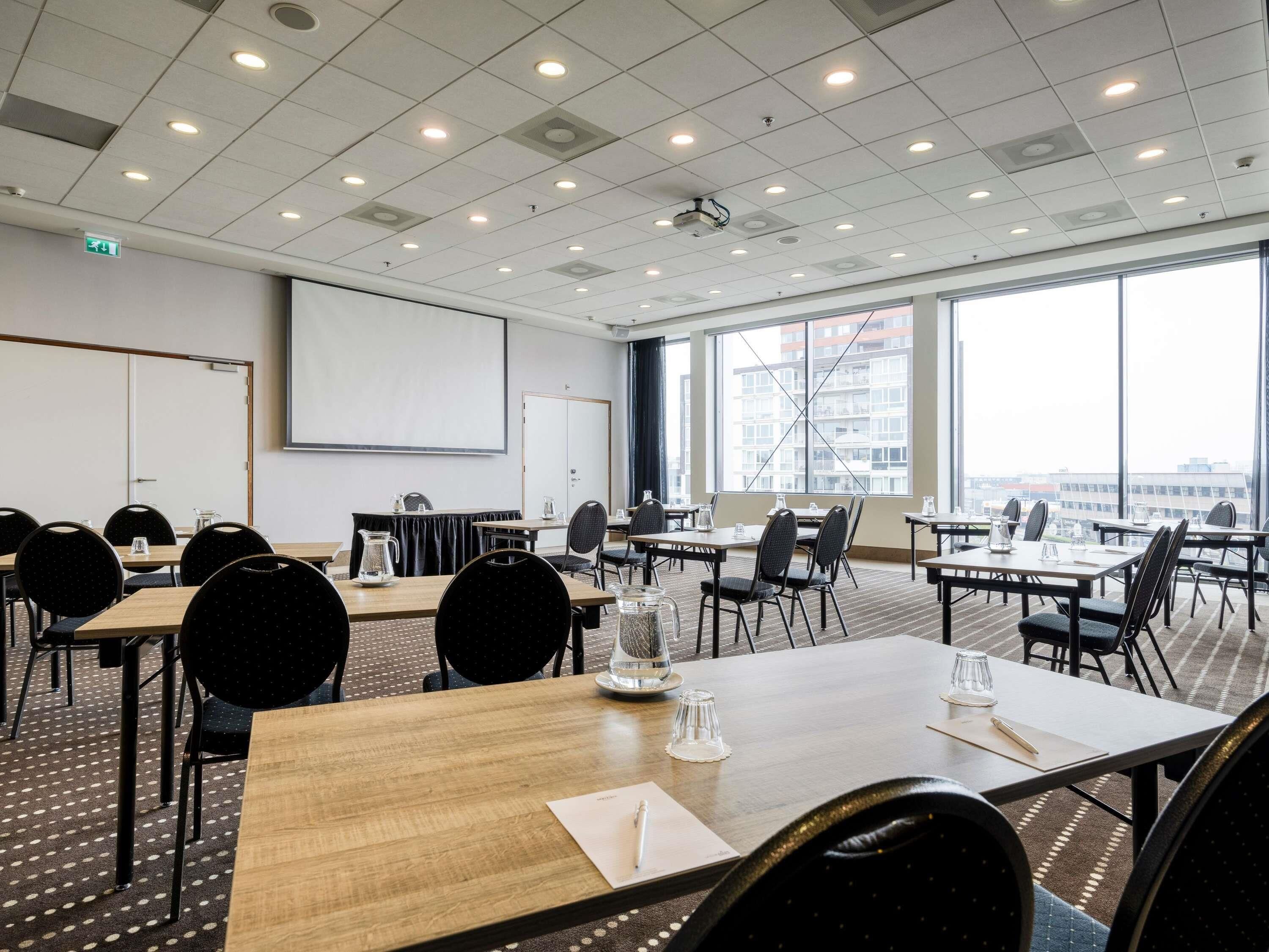 A conference room with multiple tables and chairs, a projector screen, and large windows offering a city view.