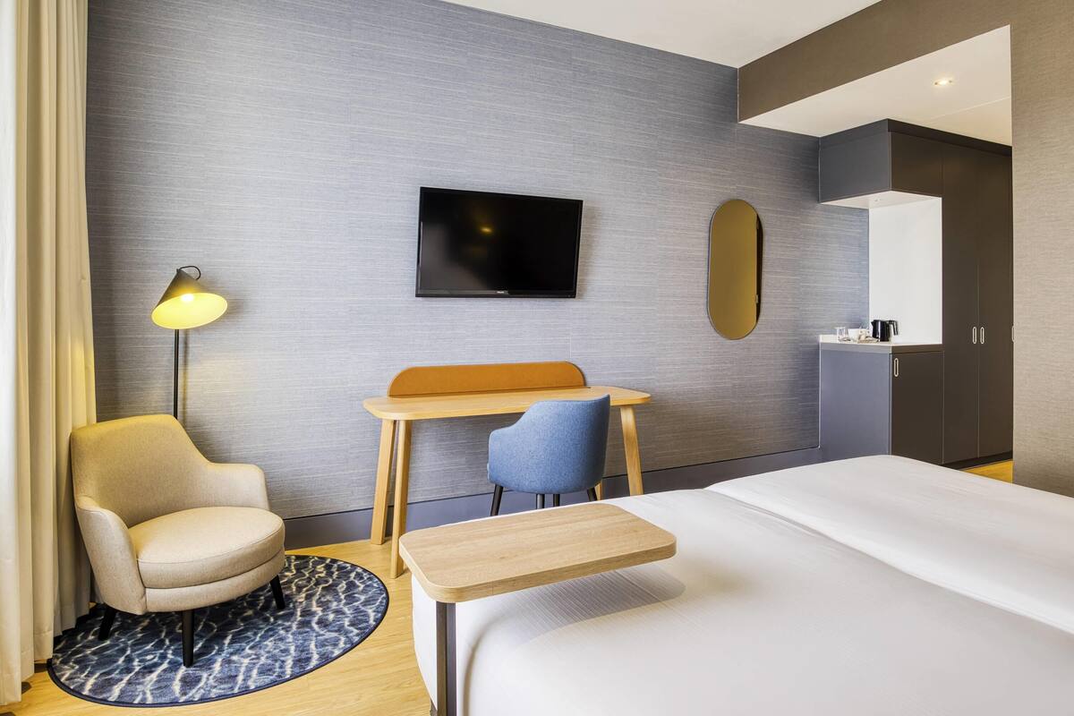 A modern hotel room with a bed, a desk, a chair, a TV, and a mirror.
