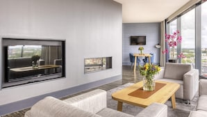 A modern living room with a flat-screen TV mounted on the wall, a wooden coffee table, grey sofas, and a view of the city through large windows.