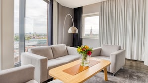 A modern hotel room with a large window, a sofa, a wooden coffee table, and a vase of flowers.