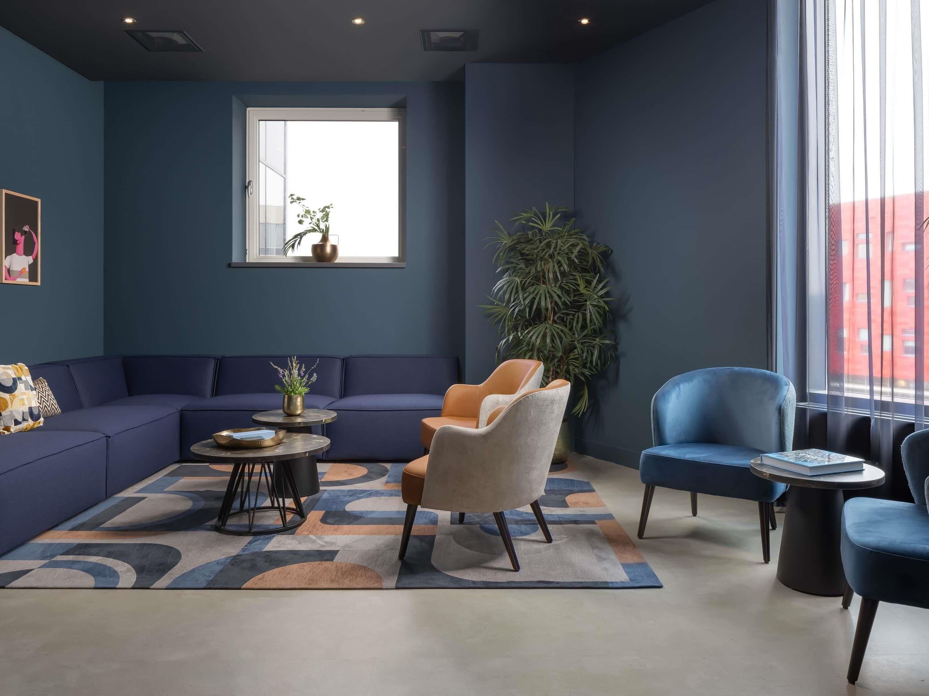 A modern living room with a blue sofa, a beige armchair, and a blue armchair. There is a large window with a view of buildings, a potted plant, and a painting on the wall.