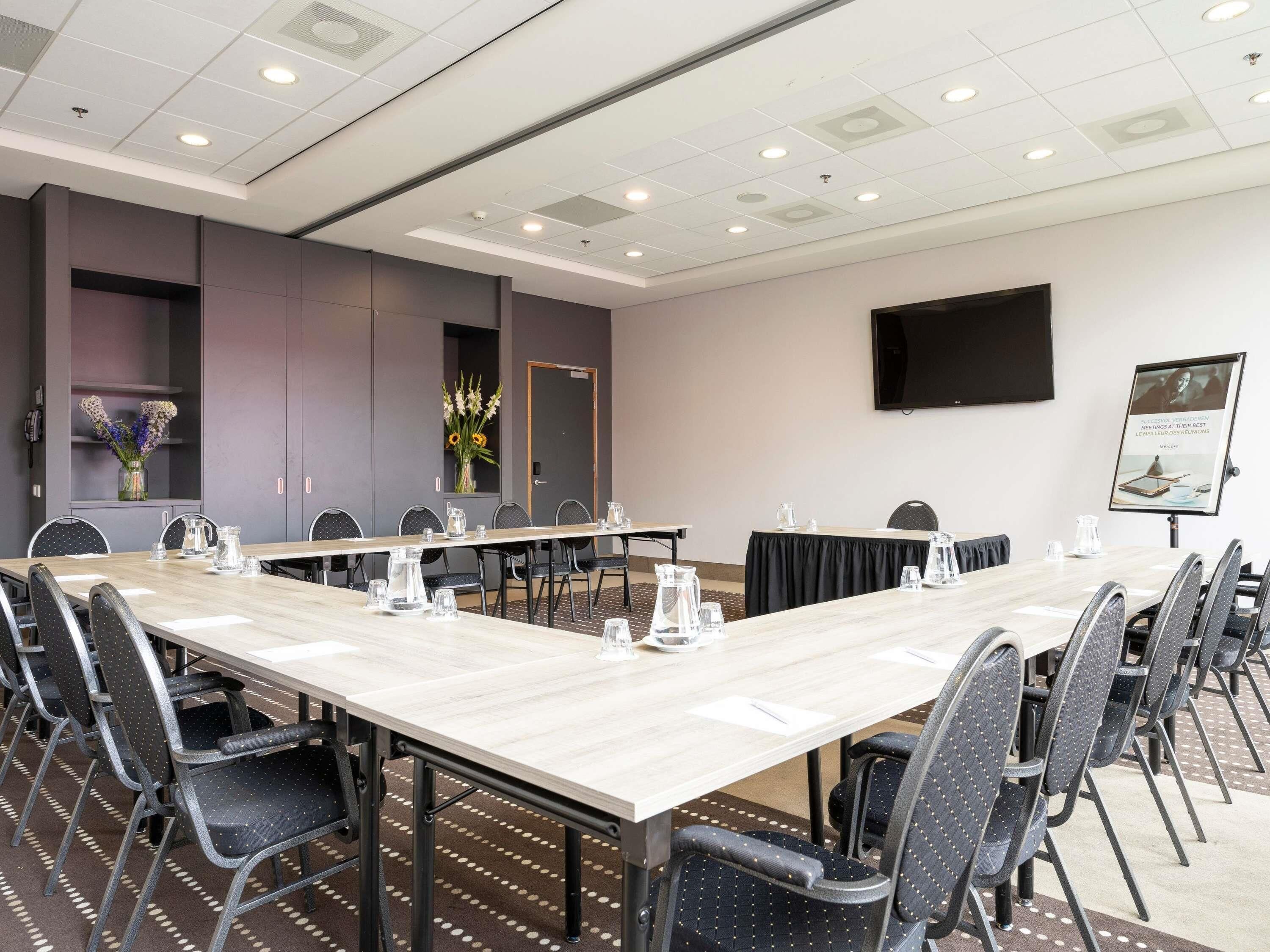 A conference room with a long table, chairs, a projector screen, and a wall-mounted TV.