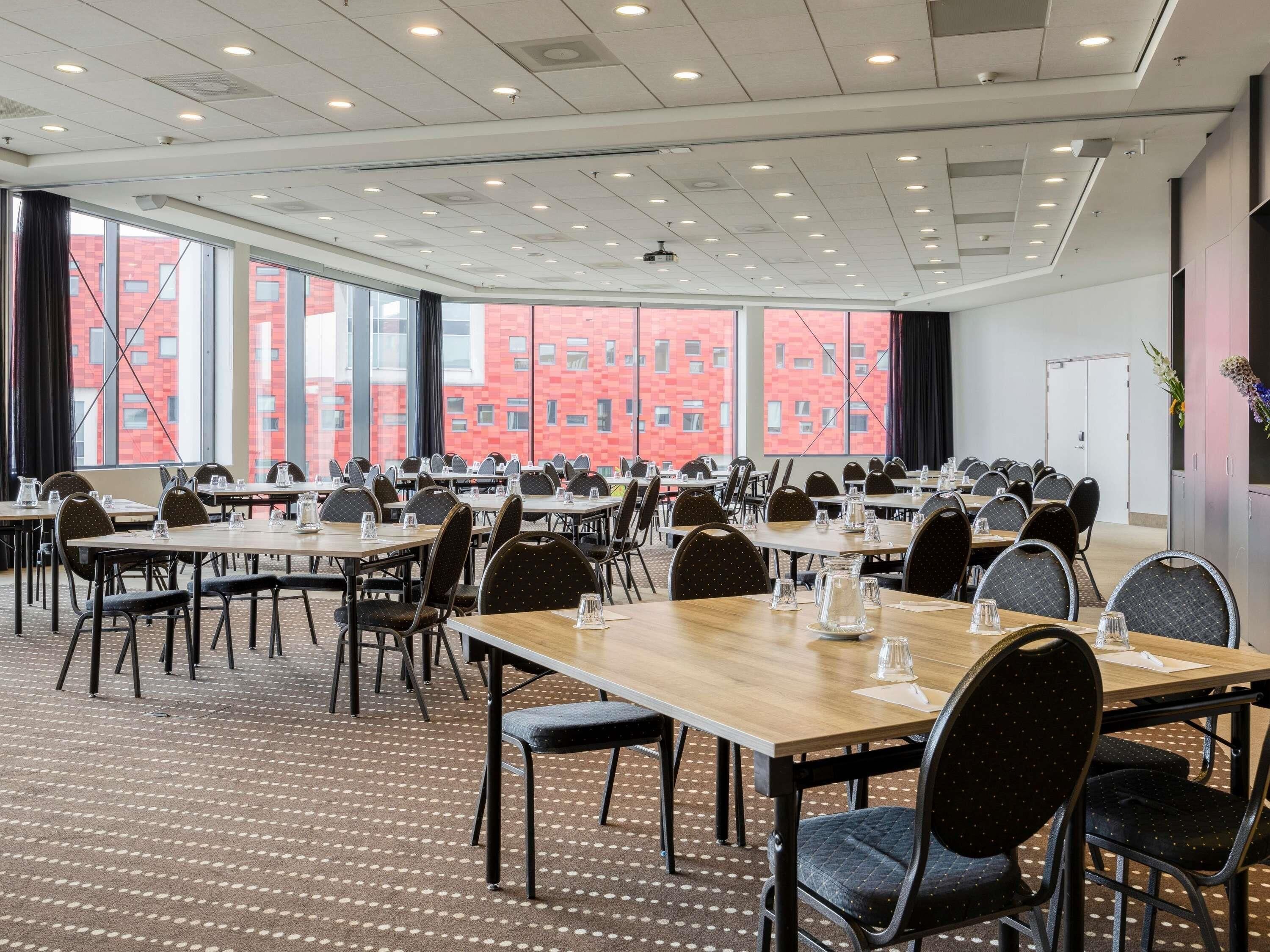Una sala conferenze con diversi tavoli e sedie, ampie finestre che offrono vista su un edificio in mattoni rossi e un soffitto con illuminazione a incasso.