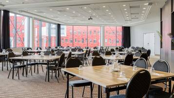 Una sala conferenze con diversi tavoli e sedie, ampie finestre che offrono vista su un edificio in mattoni rossi e un soffitto con illuminazione a incasso.