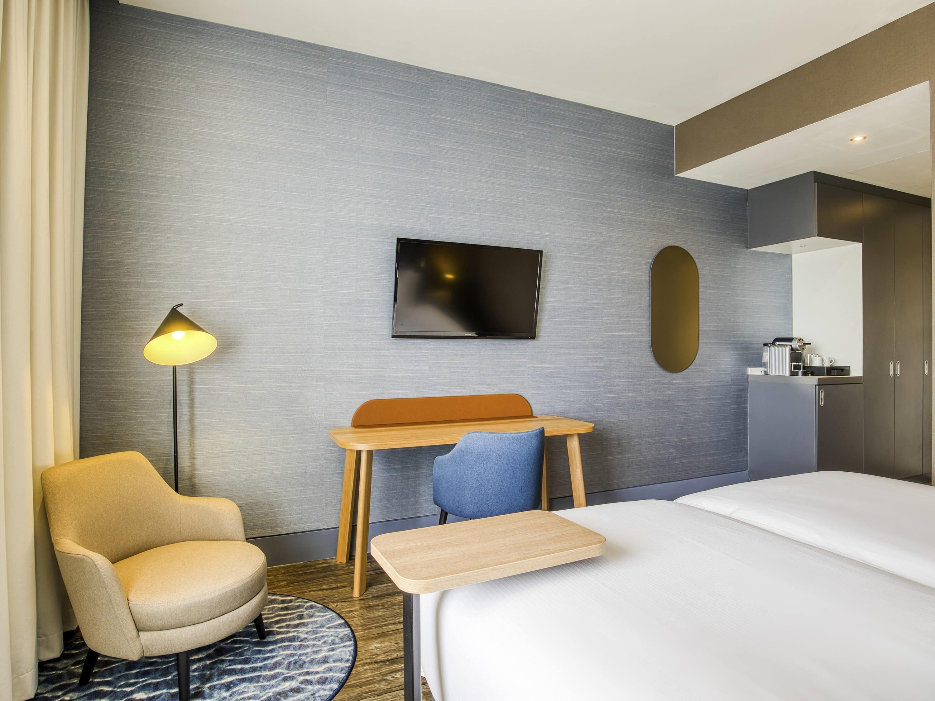 Habitación de hotel con cama, escritorio, silla, televisor, espejo y cafetera.