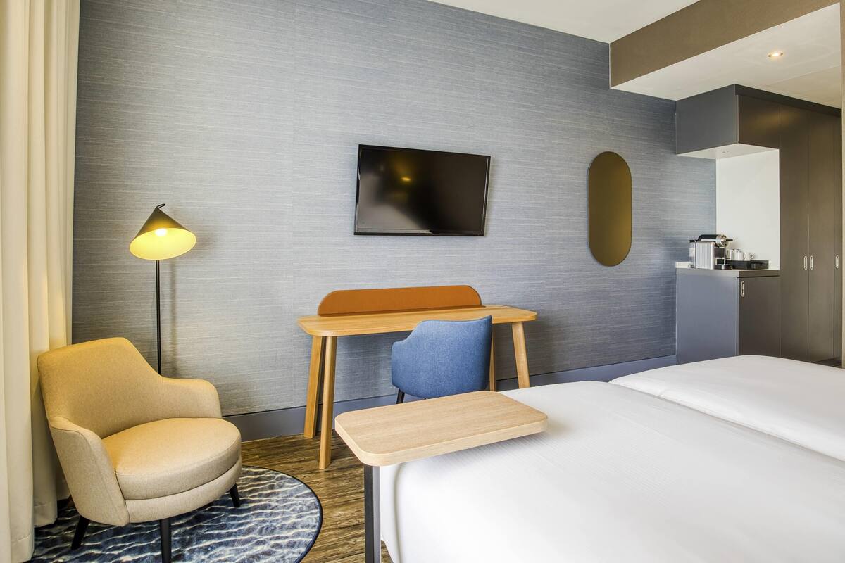 A hotel room with a bed, a desk, a chair, a TV, a mirror, and a coffee machine.