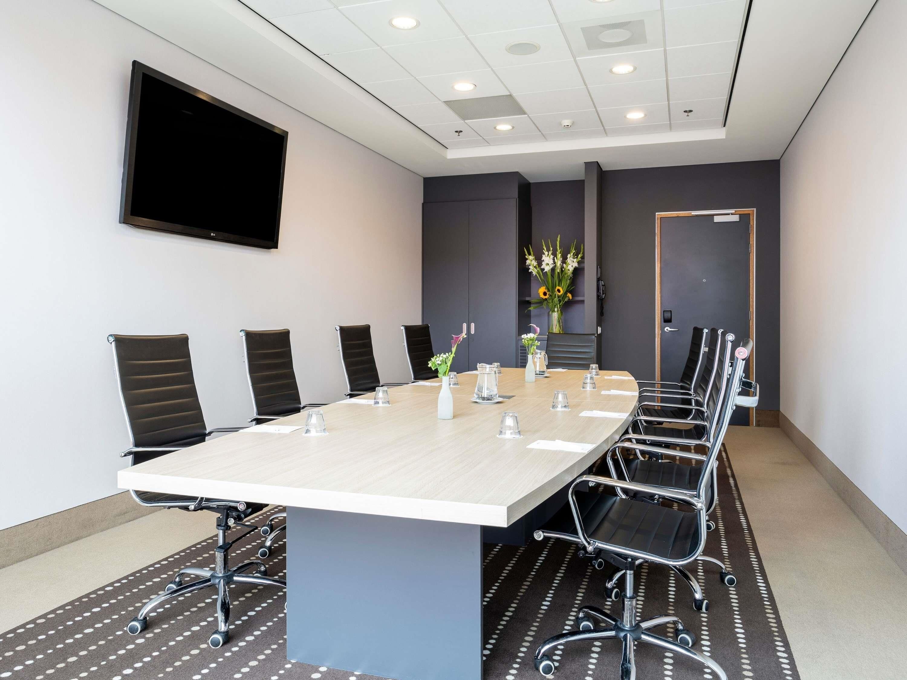 A conference room with a long table, black chairs, a flat-screen TV, and a vase with flowers.
