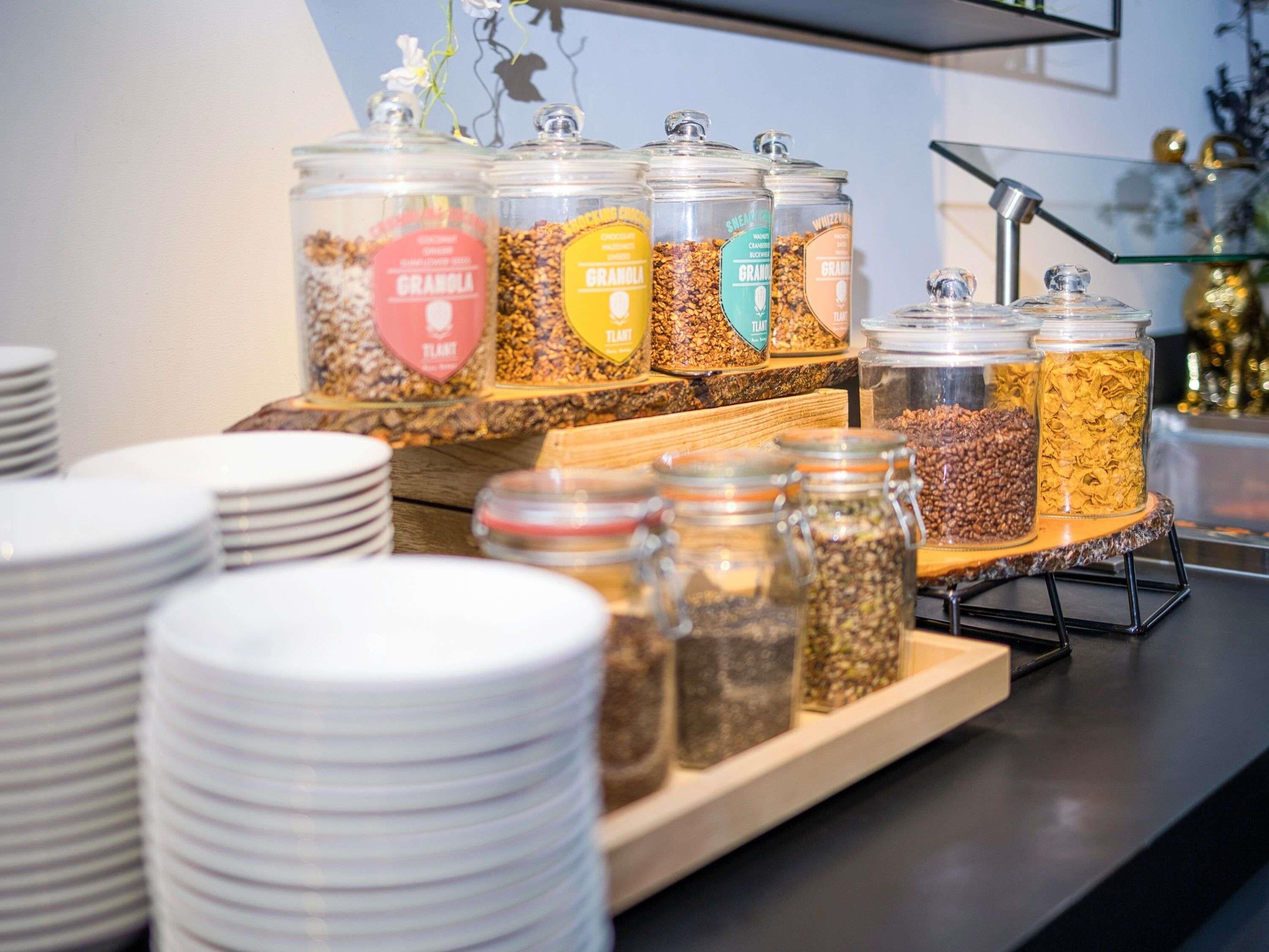 Un tavolo da buffet con diversi cibi esposti in contenitori di vetro, una pila di piatti bianchi e un supporto metallico.