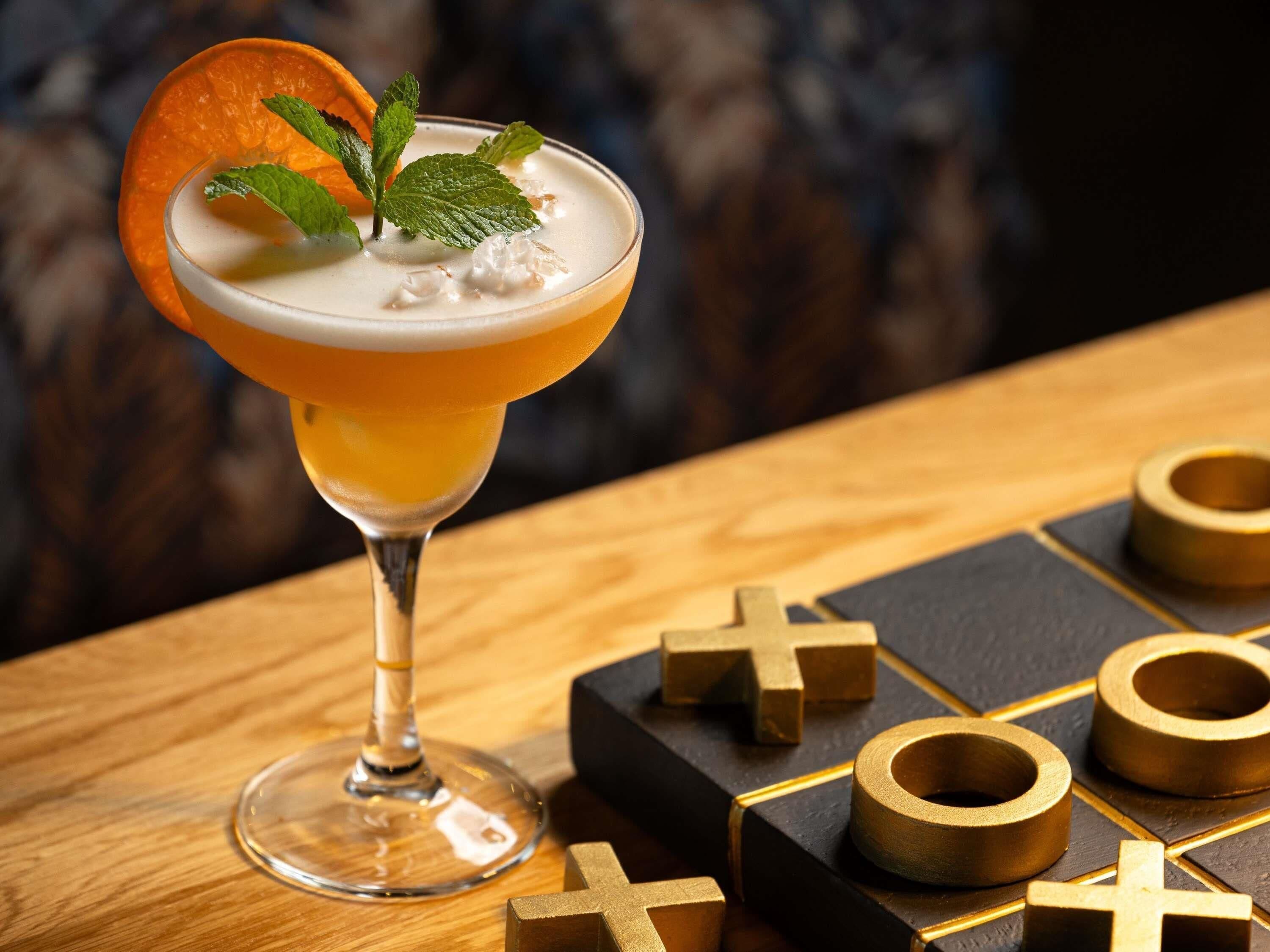 A cocktail in a glass with a slice of orange and mint garnish, placed on a wooden table with a chessboard and pieces.