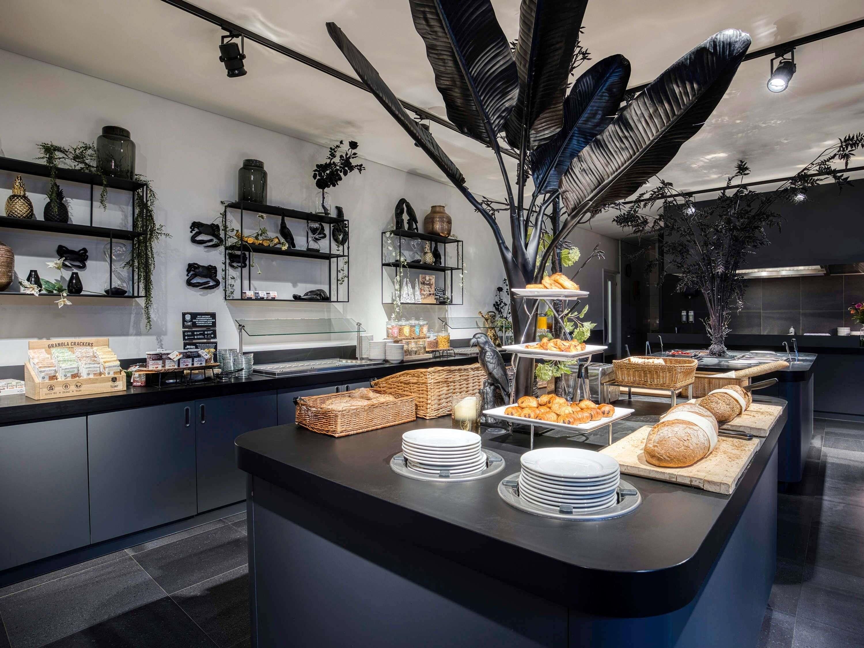 Zona de bufé con diversos alimentos, incluyendo pan y pastelería, y elementos decorativos como estructuras similares a plumas negras y estanterías con plantas y adornos.