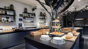 Zona de bufé con diversos alimentos, incluyendo pan y pastelería, y elementos decorativos como estructuras similares a plumas negras y estanterías con plantas y adornos.