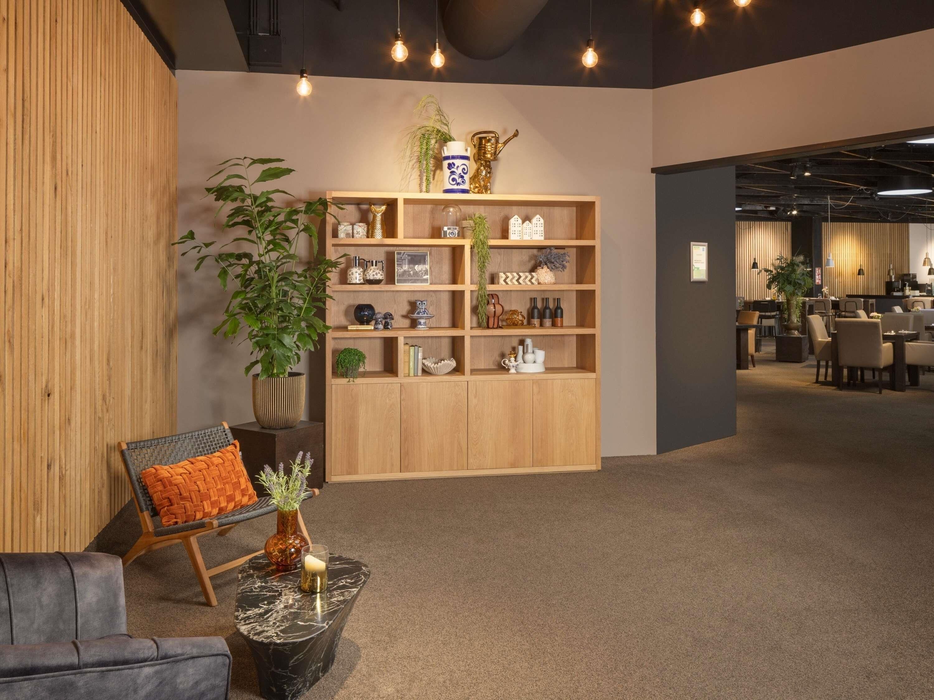 A modern interior with a wooden bookshelf, a seating area with a chair and a table, and a dining area in the background.