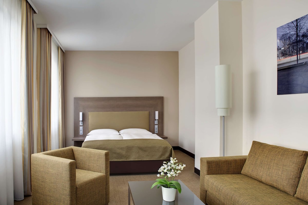 Une chambre d’hôtel avec un lit, deux fauteuils, une table basse avec une plante, une lampe et un tableau au mur.