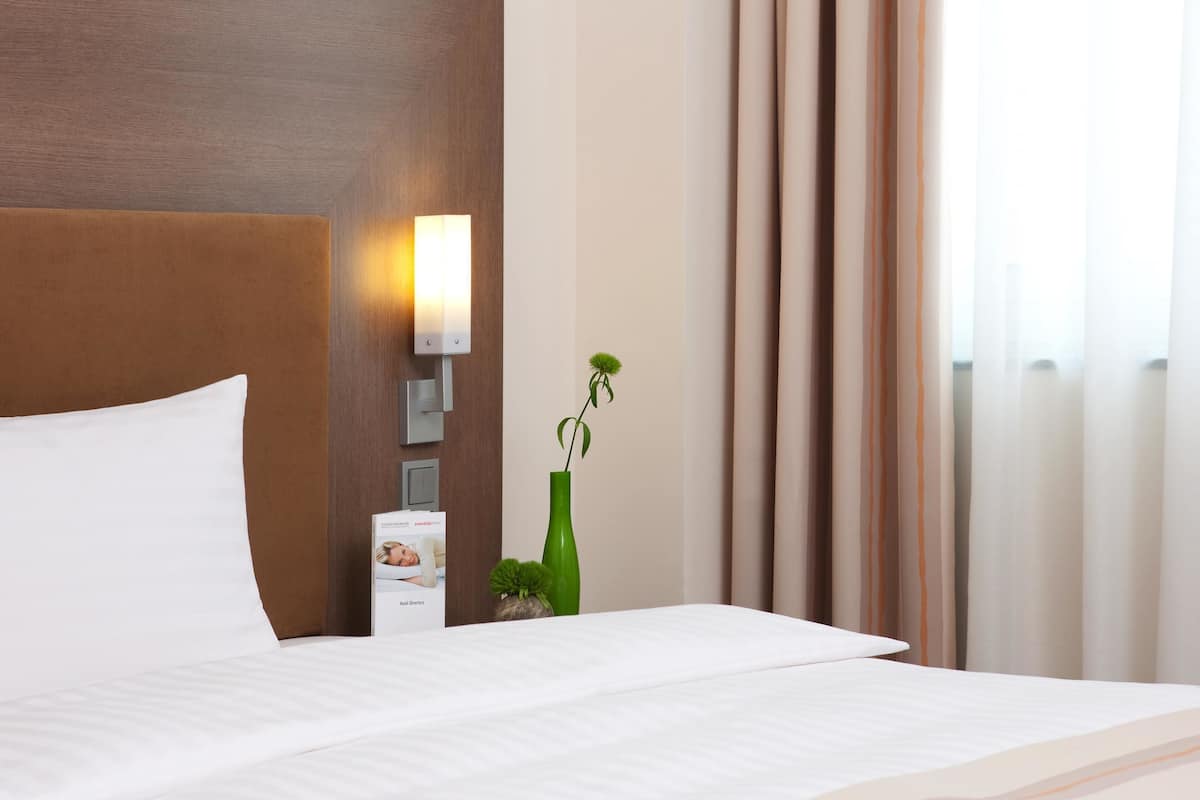 Une chambre d’hôtel avec un grand lit, une table de chevet avec une lampe, une petite plante verte et une fenêtre avec des rideaux.