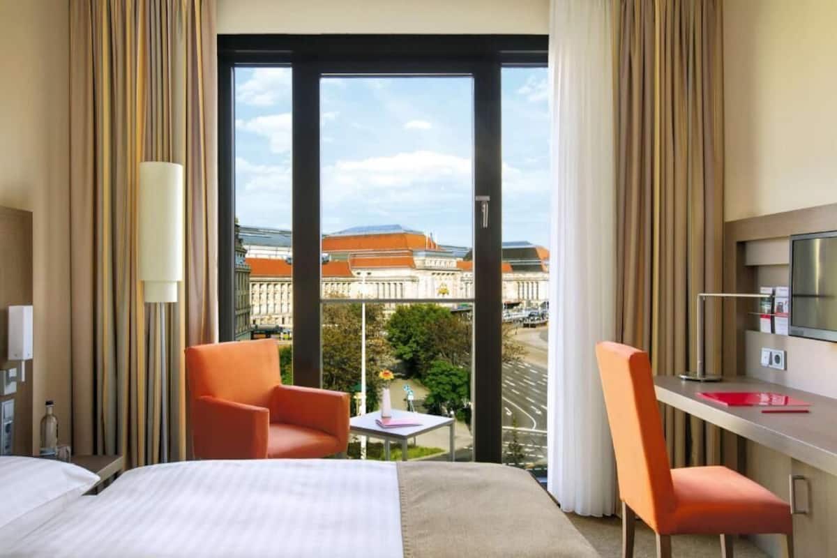Une chambre d’hôtel avec un lit, deux fauteuils orange, un bureau avec une télévision et une grande fenêtre donnant sur la ville.