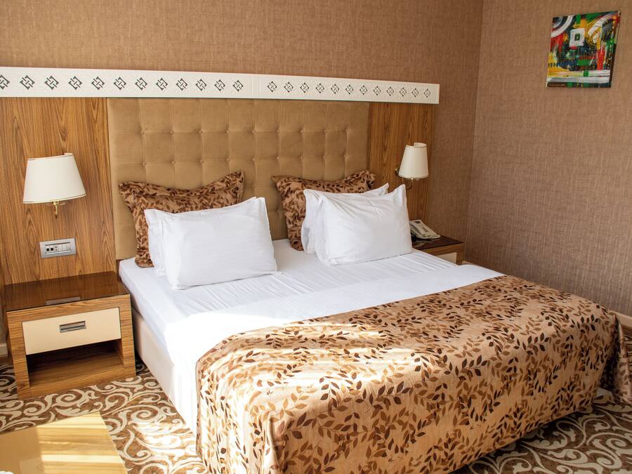 Quarto de hotel com cabeceira estofada, duas luminárias de cabeceira, cama com roupa de cama branca e manta decorativa.
