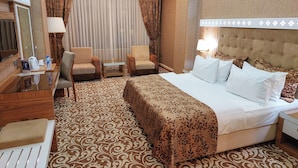 Habitación de hotel con una cama grande, dos sillones, una mesita de noche con lámpara, televisión y alfombra estampada.