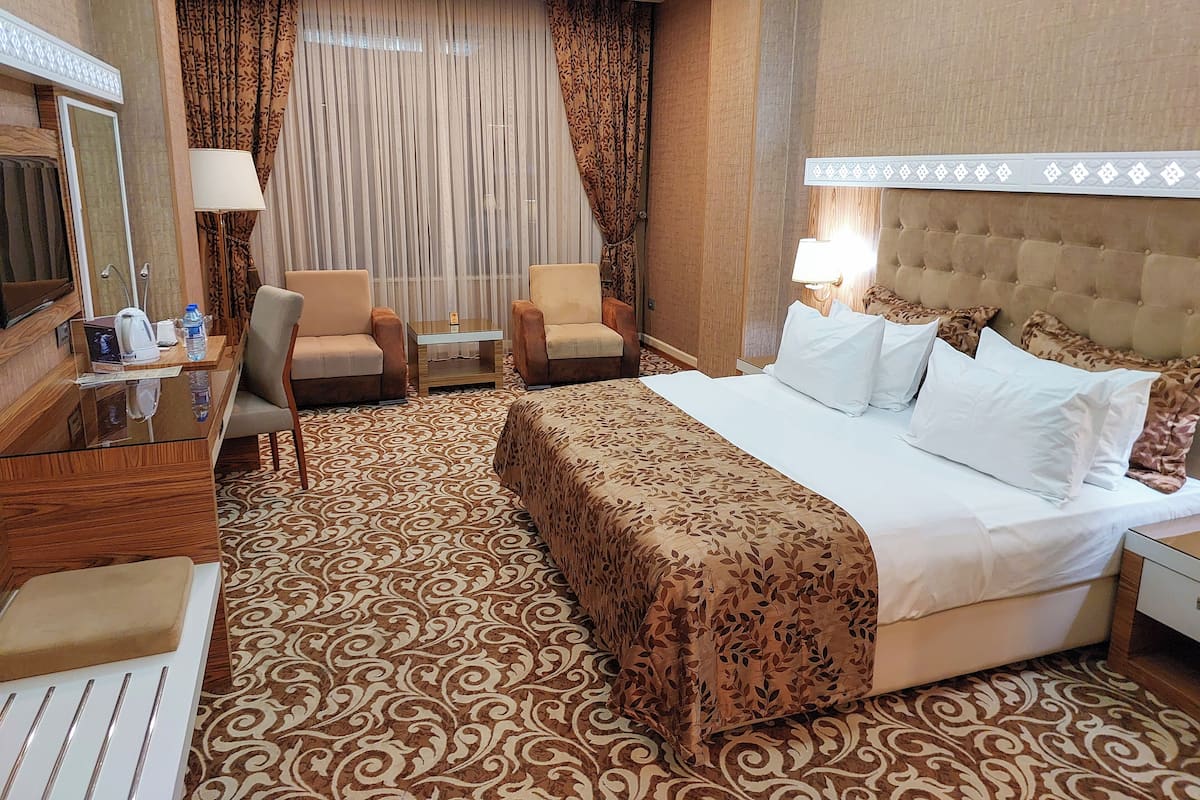 A hotel room with a large bed, two armchairs, a small table with a lamp, a TV, and a patterned carpet.
