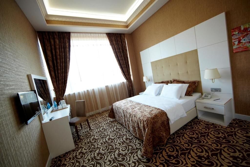 A hotel room with a bed, bedside tables, a desk, a television, and a patterned carpet.