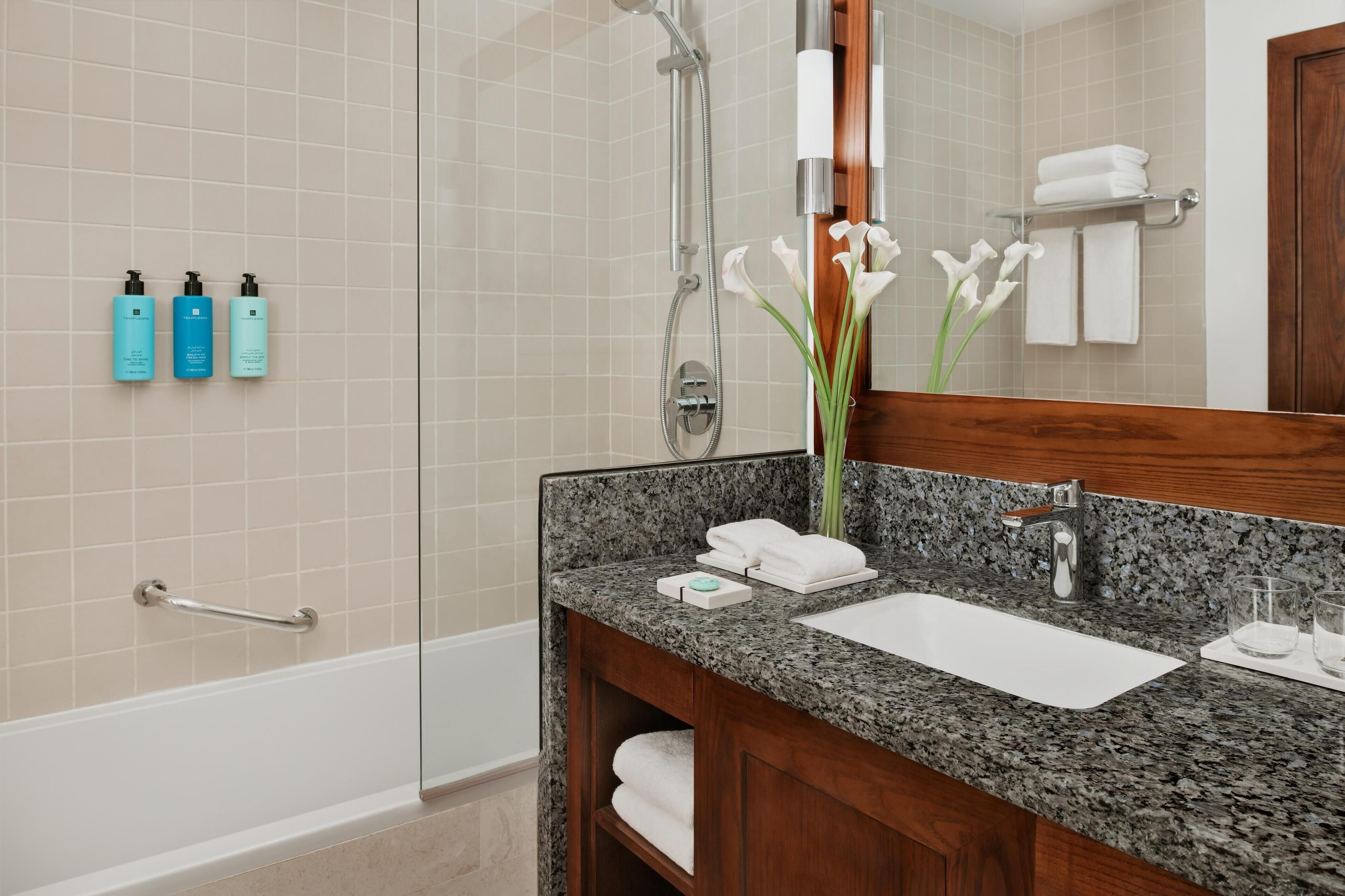 Un baño moderno con encimera de granito, lavamanos, espejo y zona de ducha.