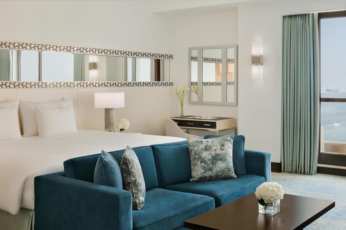 A hotel room with a large bed, a blue sofa, a small table with a vase of flowers, a desk with a lamp, and a view of the sea through large windows.