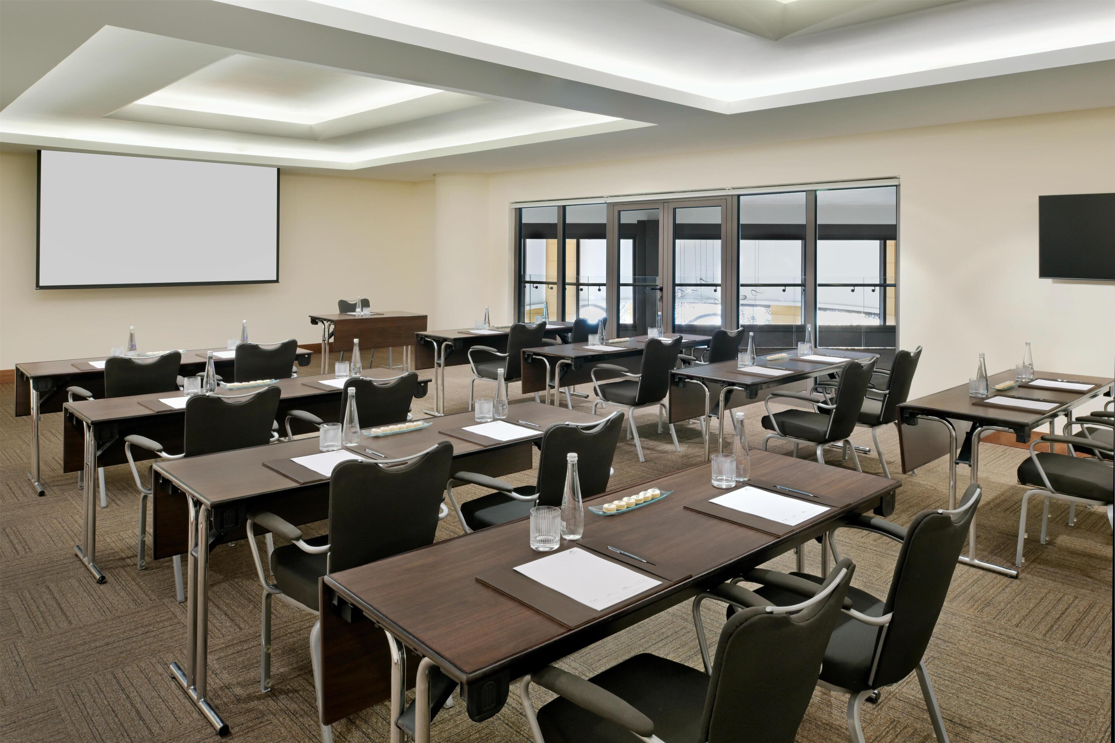 A conference room with multiple tables, chairs, a projector screen, and large windows offering a view of the outside.