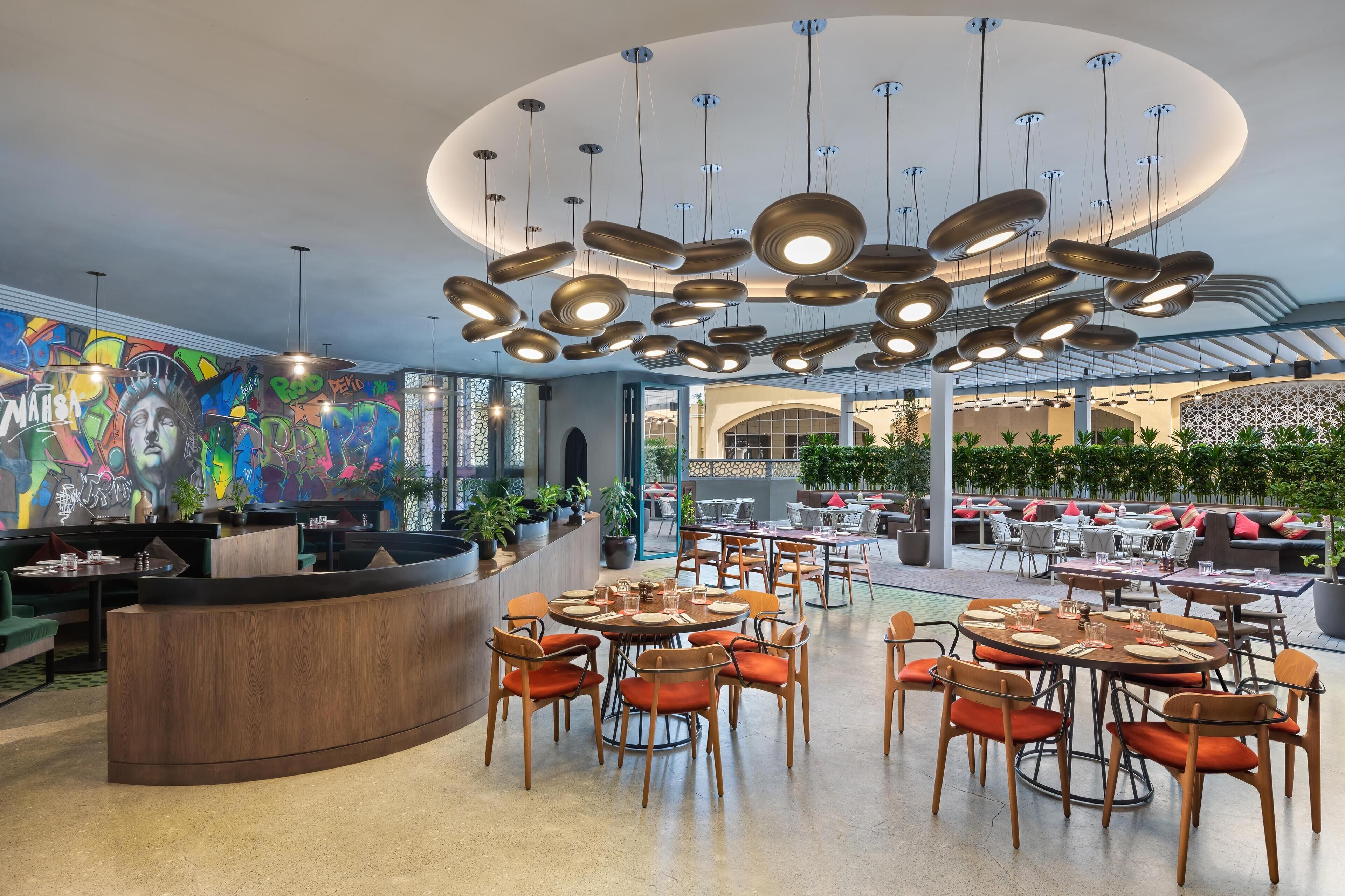 A modern restaurant with round tables and orange chairs, a colorful graffiti wall, and a ceiling with circular light fixtures.