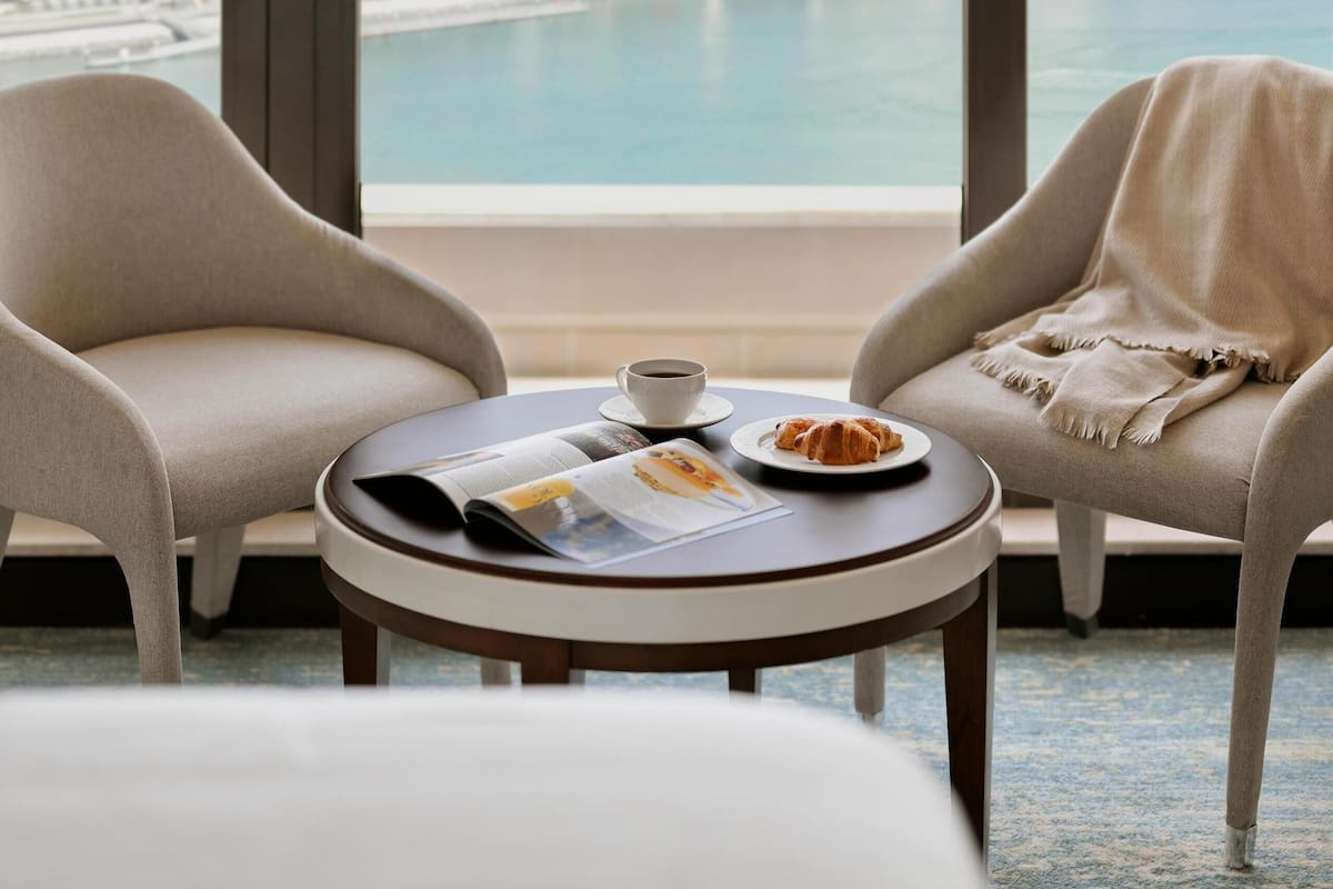 A hotel room with two armchairs, a small table with a croissant and a cup, and a view of a Ferris wheel and water.