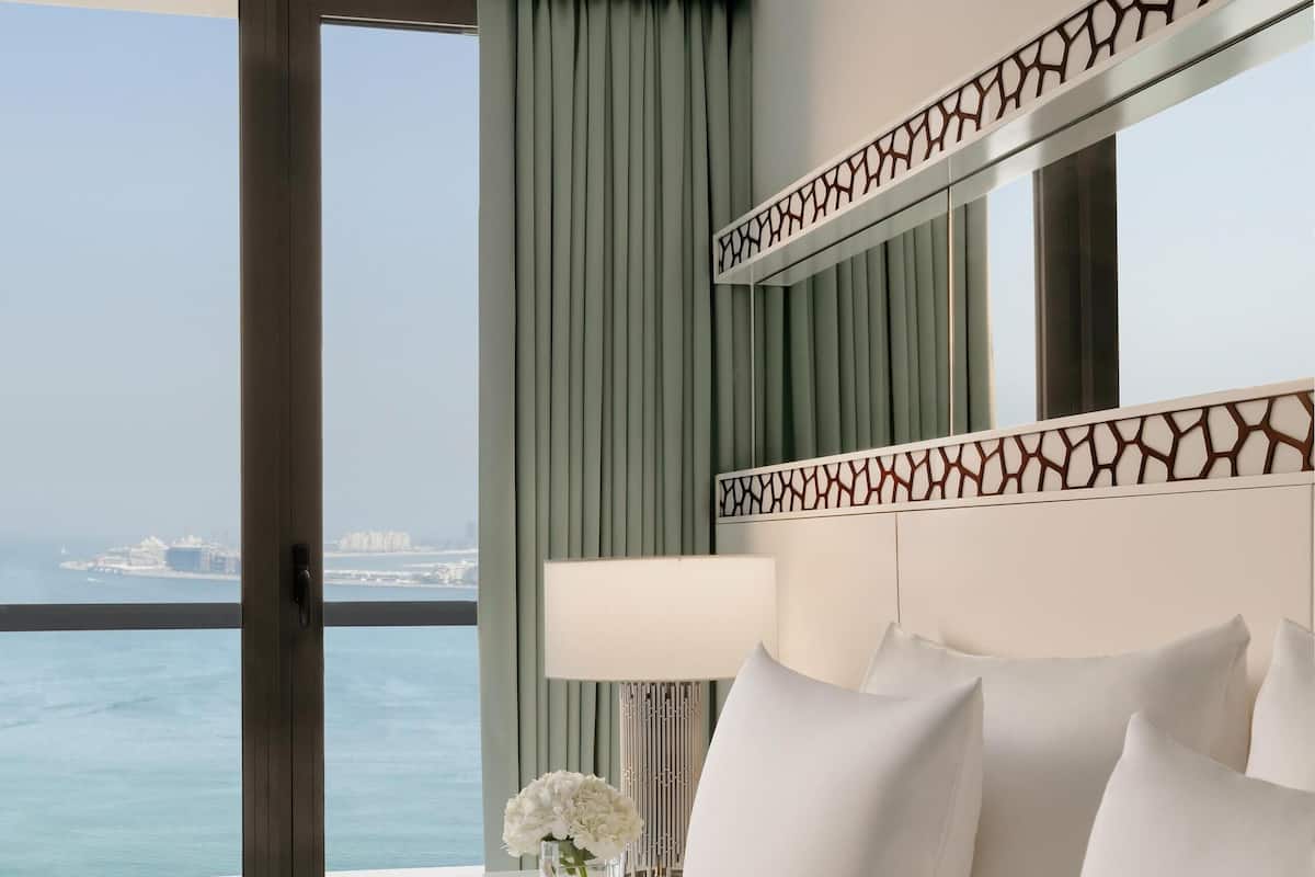 A hotel room with a large bed, a view of the sea, a decorative headboard, and a nightstand with a lamp and flowers.