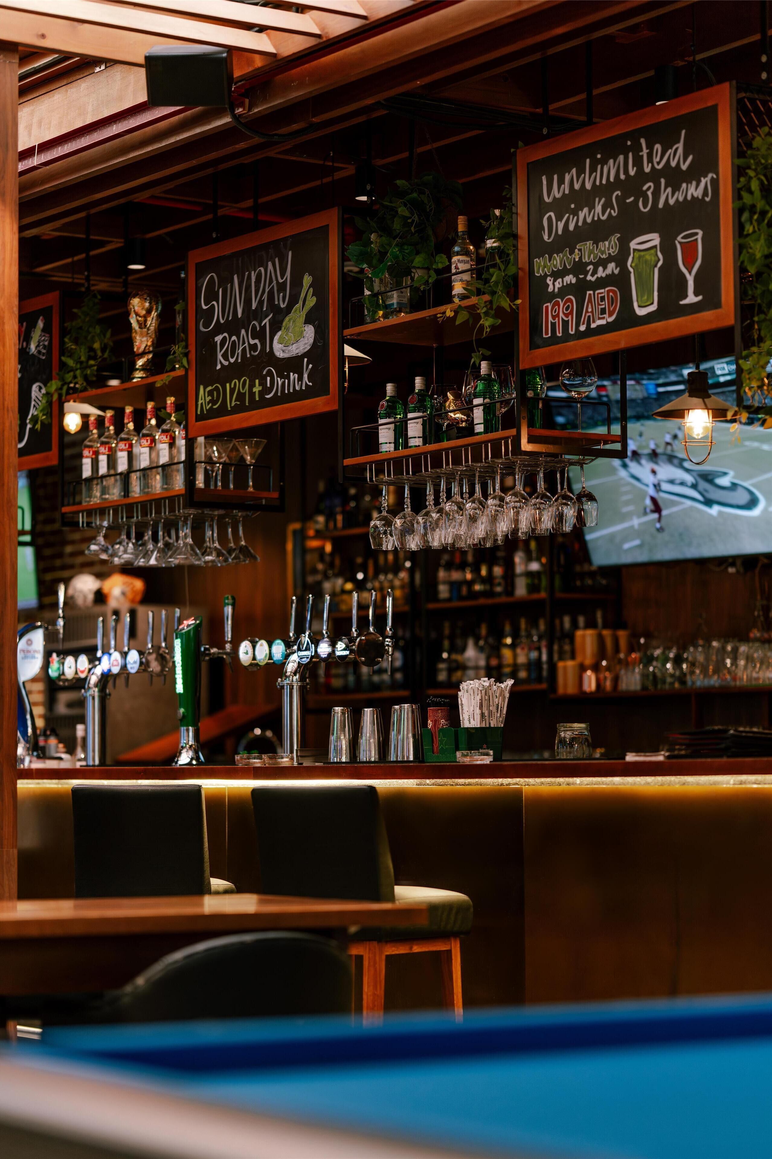 Un espace bar avec une affiche proposant des boissons à volonté pendant 3 heures, un menu spécial pour le dimanche avec un plat rôti, et un prix de 199 AED.