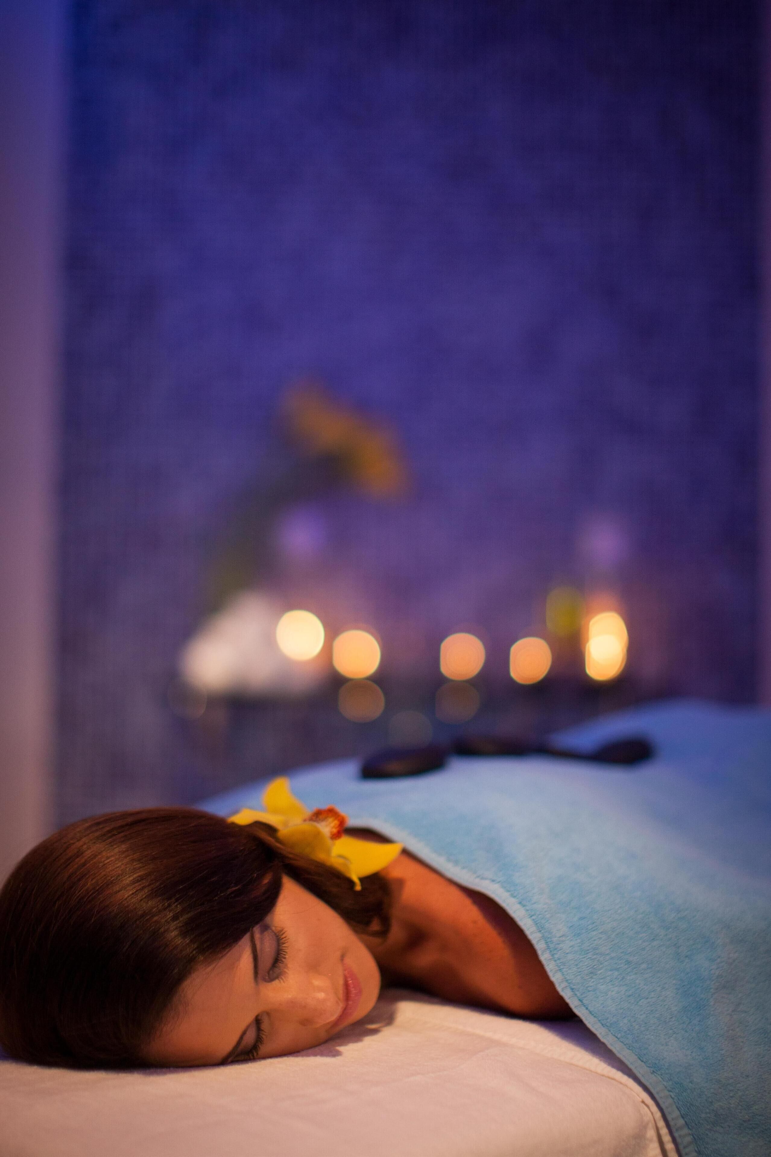 A person lying on a bed with a blue towel, a yellow flower, and candles in the background.