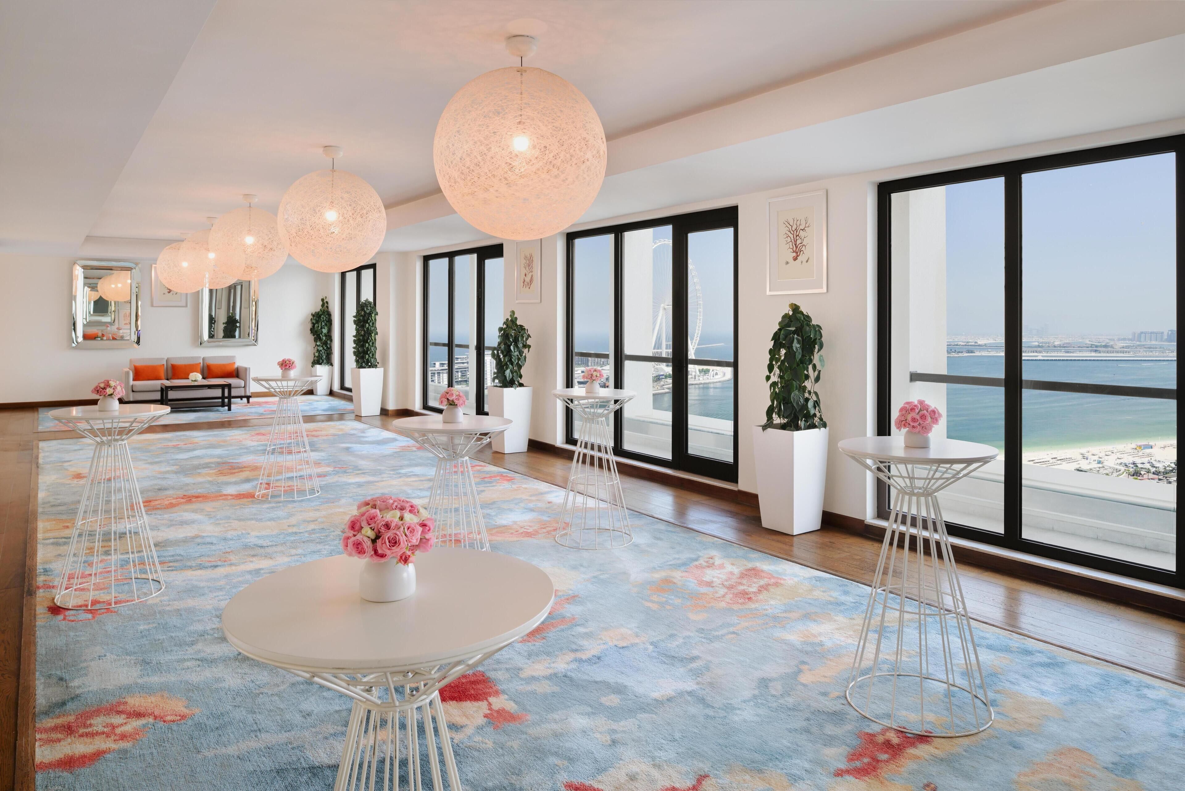 A spacious hotel room with large windows, a floral carpet, and modern furniture.