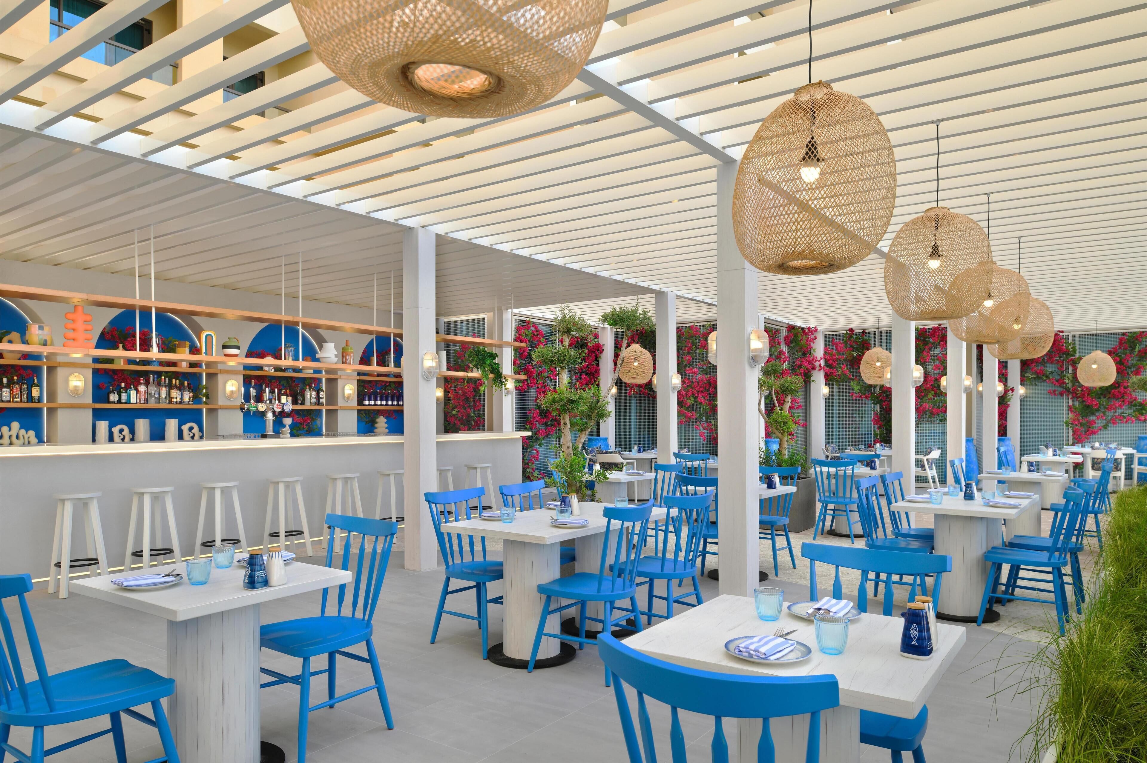 A restaurant with blue chairs and white tables, a bar area with shelves of bottles, and hanging wicker lamps.