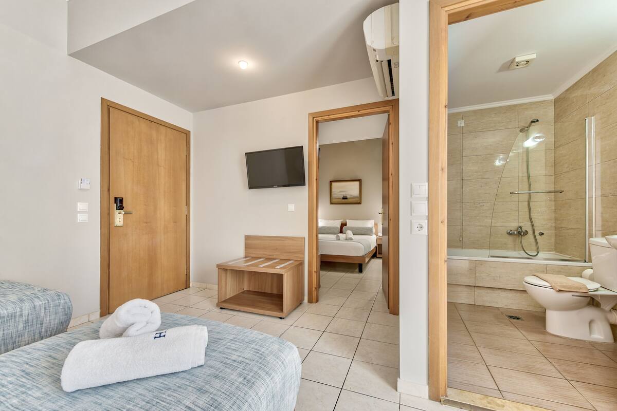 Una habitación de hotel moderna con una puerta de madera, una cama con colcha a cuadros azul y blanco, un pequeño banco de madera, un televisor montado en la pared y un baño con cabina de ducha de vidrio.