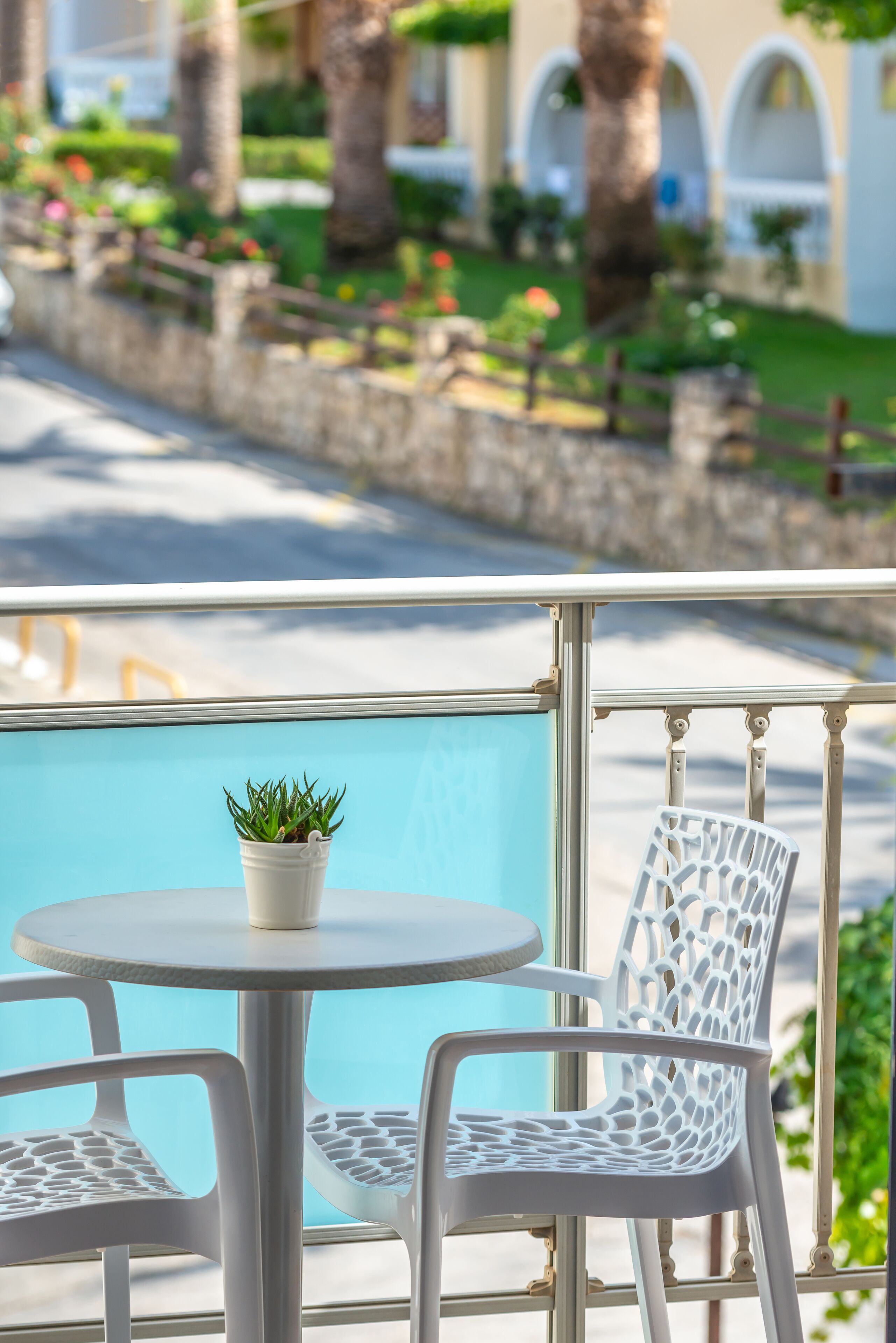 A balcony with a round table and two chairs, a potted plant, and a view of a pool and greenery.
