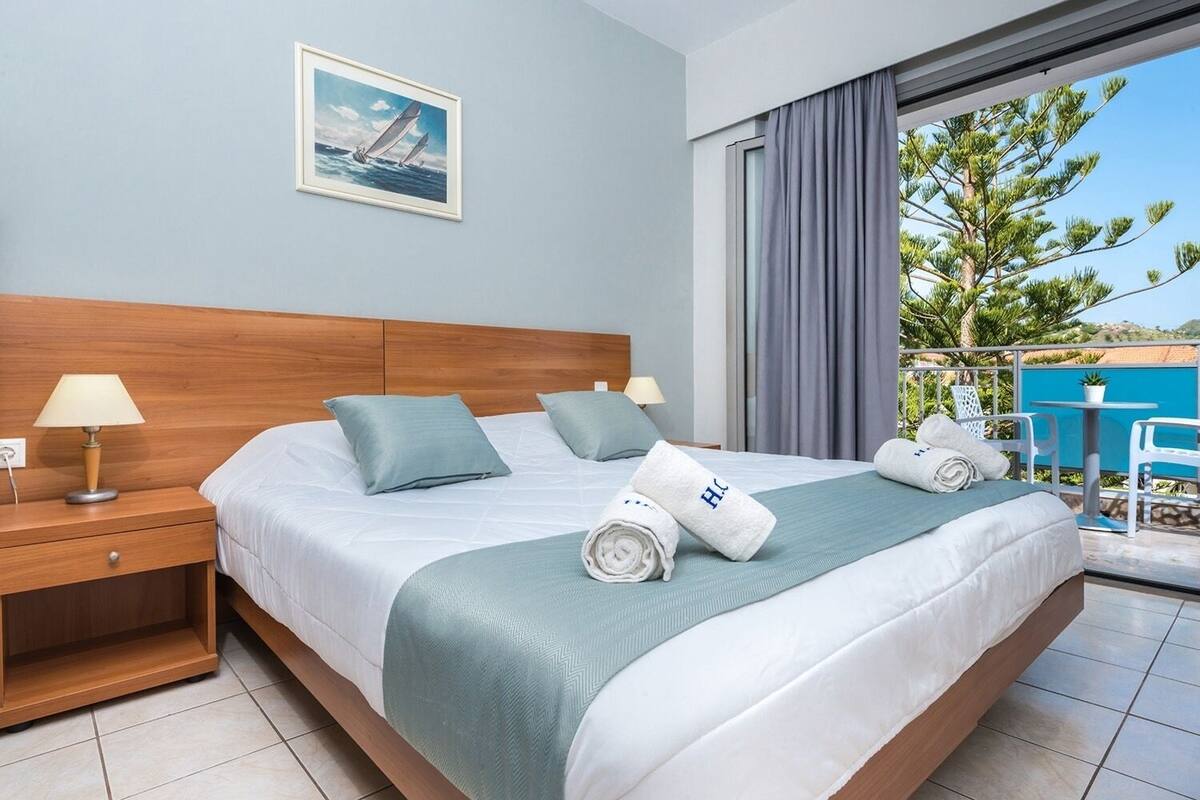 Habitación de hotel con una cama grande, una mesita de noche, una lámpara, un cuadro de veleros y un balcón con vistas a árboles y un edificio azul.