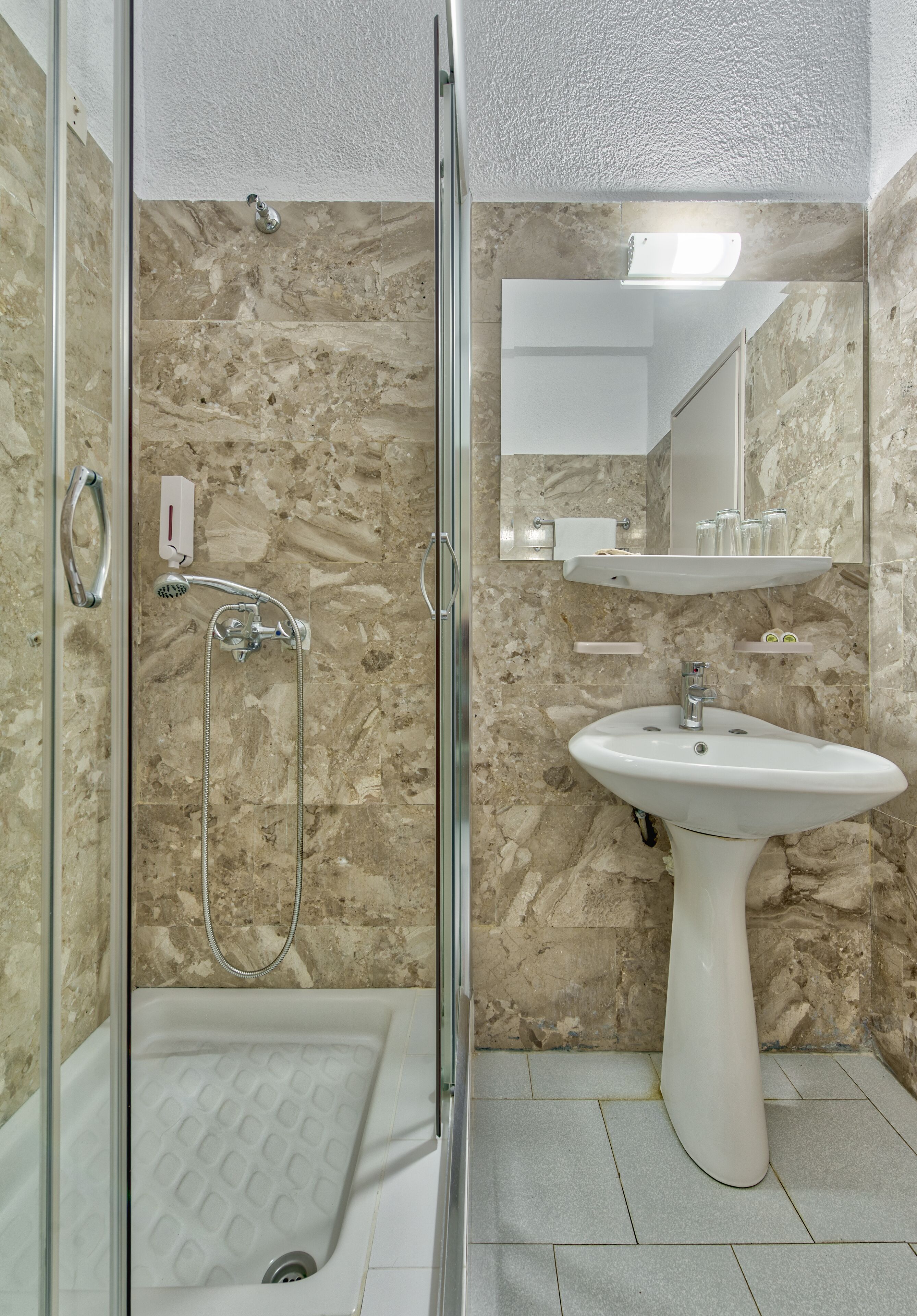 A bathroom with a glass-enclosed shower, a pedestal sink, and a marble-tiled wall.