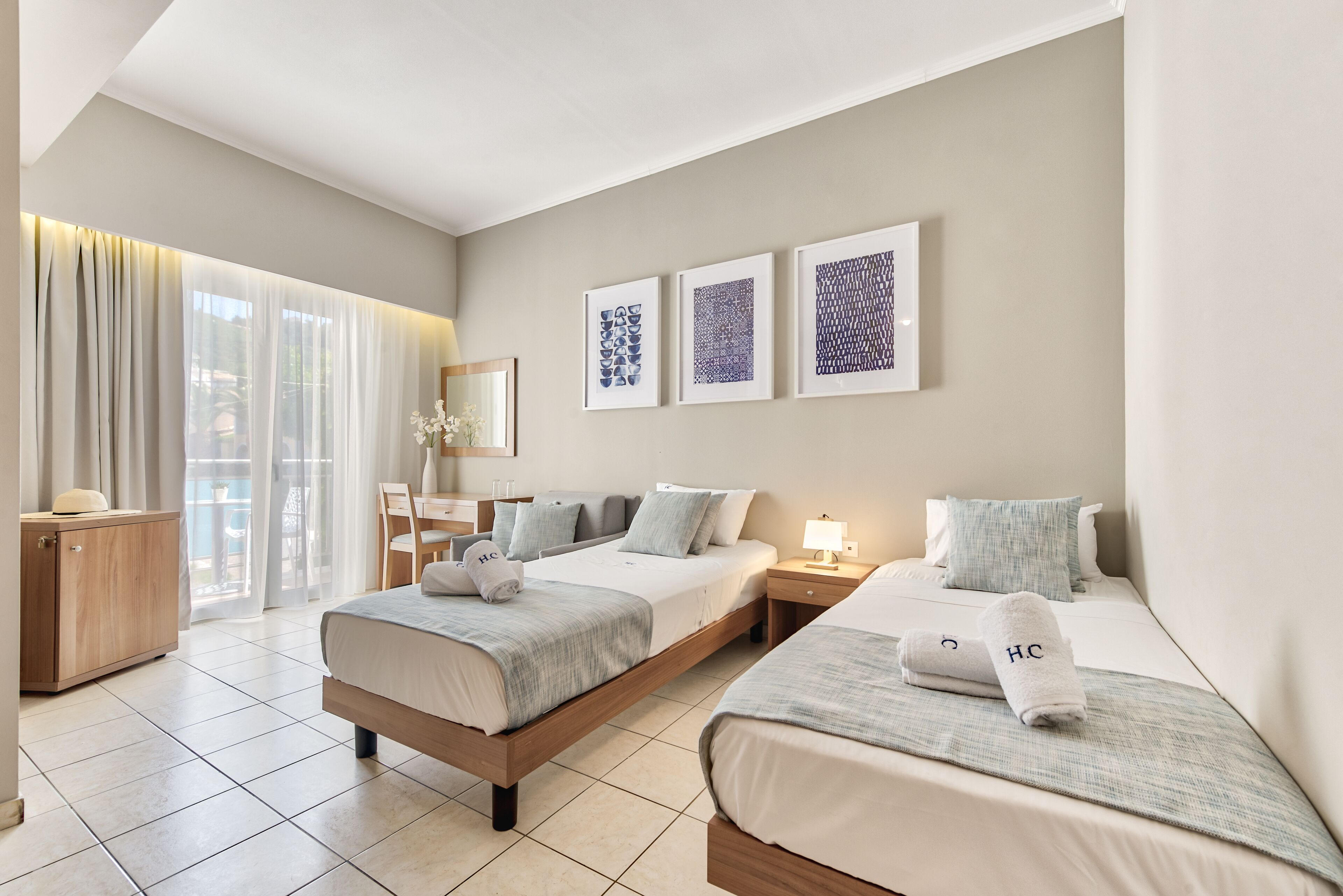A hotel room with two beds, a desk, and a chair. There are three framed artworks on the wall.