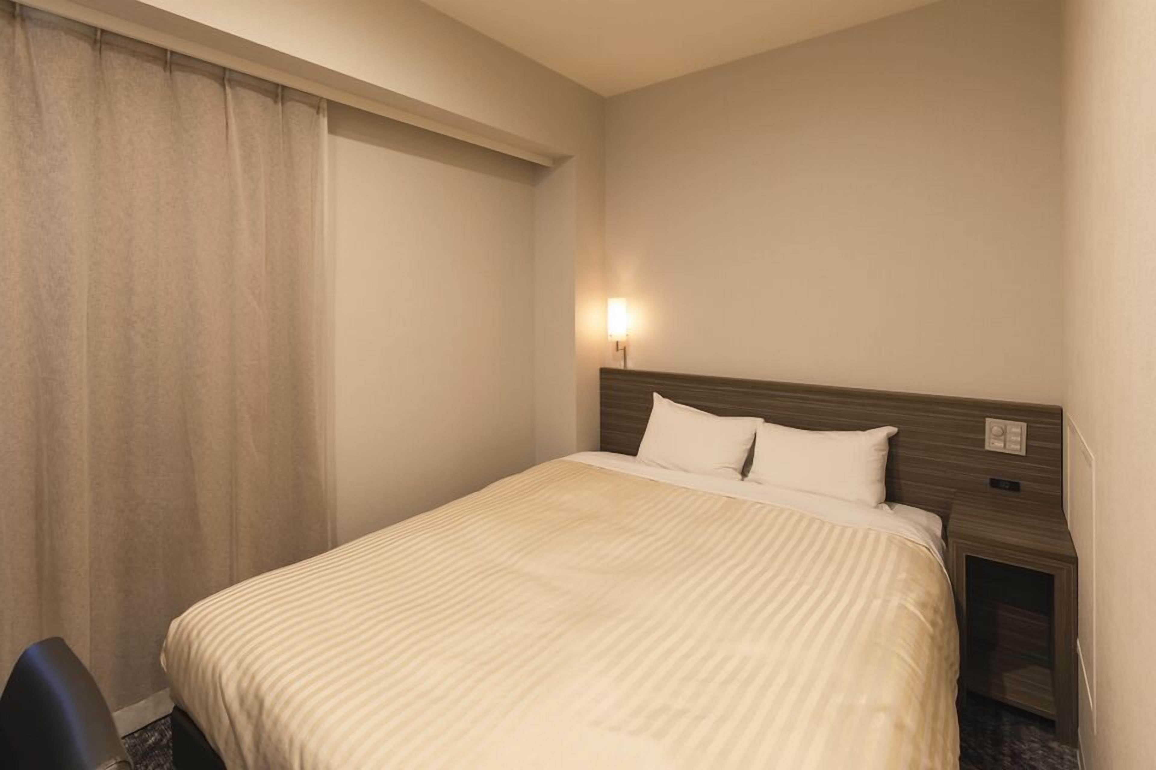 Quarto de hotel com cama, mesa de cabeceira, luminária e janela com cortinas.