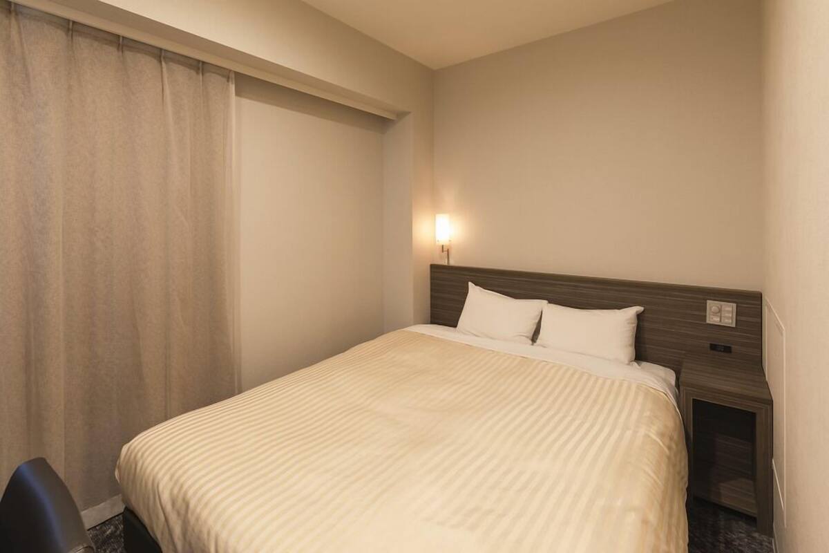 A hotel room with a bed, a nightstand, a lamp, and a window with curtains.